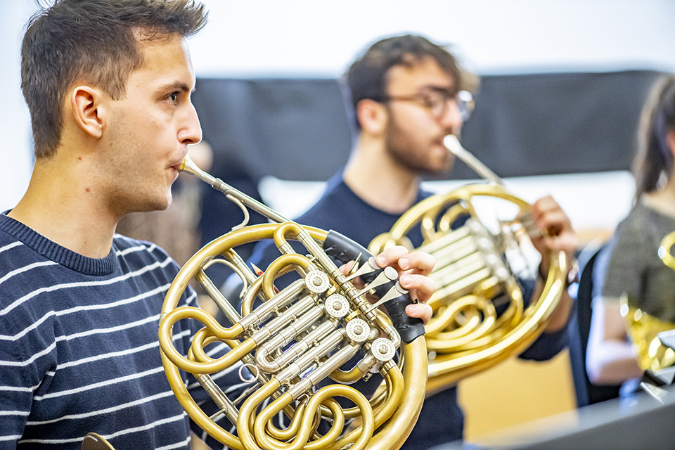 A male student performing on the French horn, with an another French hornists performing on the background.